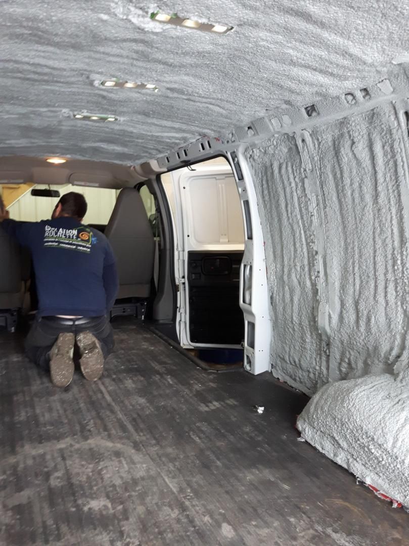 Un homme agenouillé dans une camionnette applique une isolation grise. L'intérieur de la camionnette présente des parois et un plancher gris.