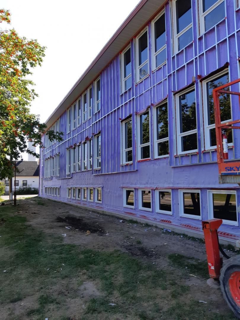 Extérieur du bâtiment en construction, montrant l'isolation violette, les fenêtres et l'ascenseur. Herbe verte et arbres.