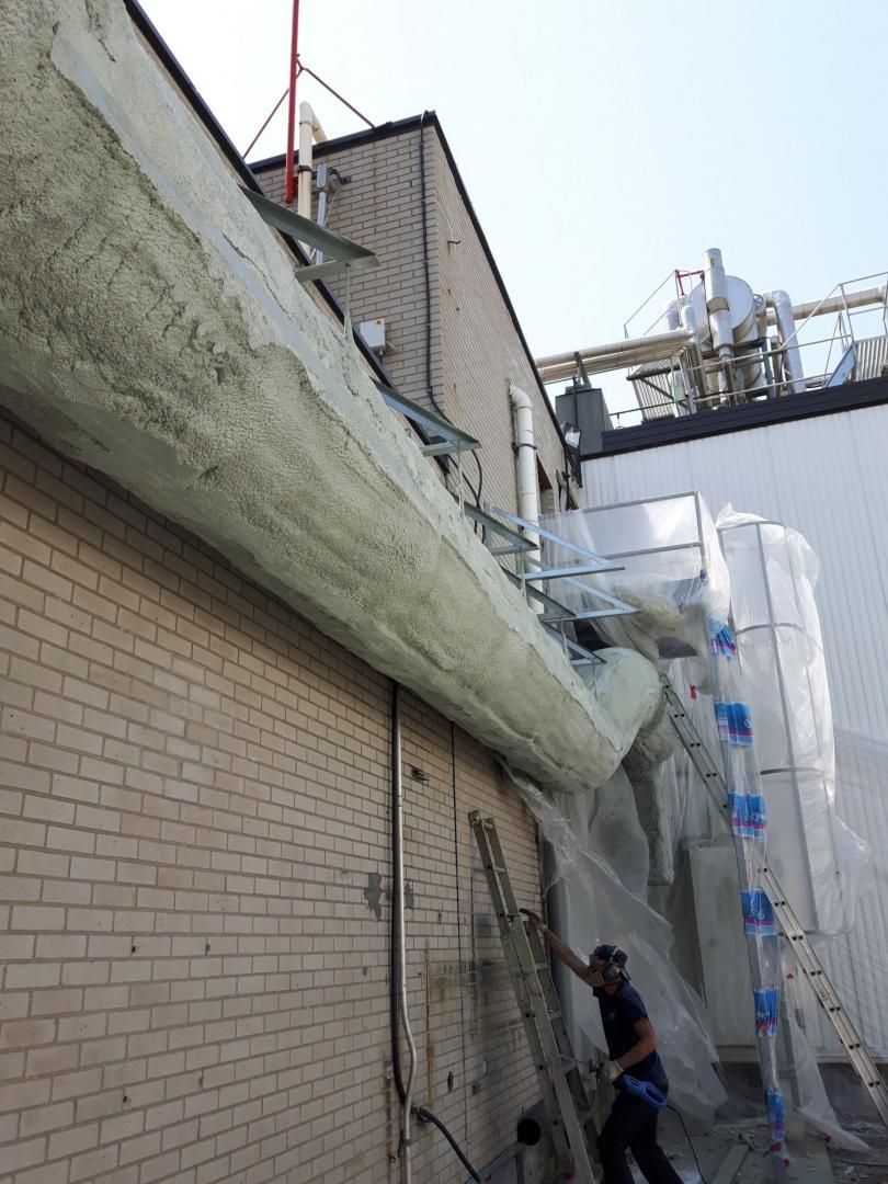 Ouvrier sur une échelle près d'un bâtiment en briques, à côté d'un gros tuyau isolé, avec des équipements industriels en arrière-plan.
