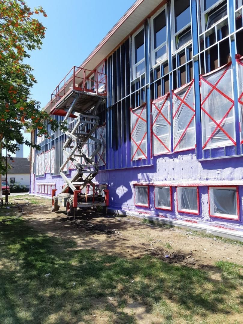 Un élévateur à ciseaux à côté d'un immeuble en construction. Revêtement violet, fenêtres bardées, journée ensoleillée.