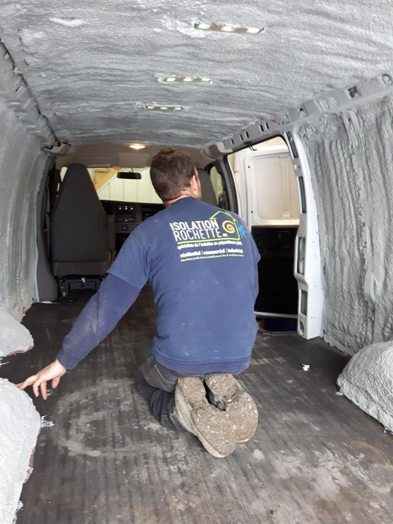 Un homme agenouillé dans une camionnette montre du doigt une mousse isolante projetée. Intérieur gris.