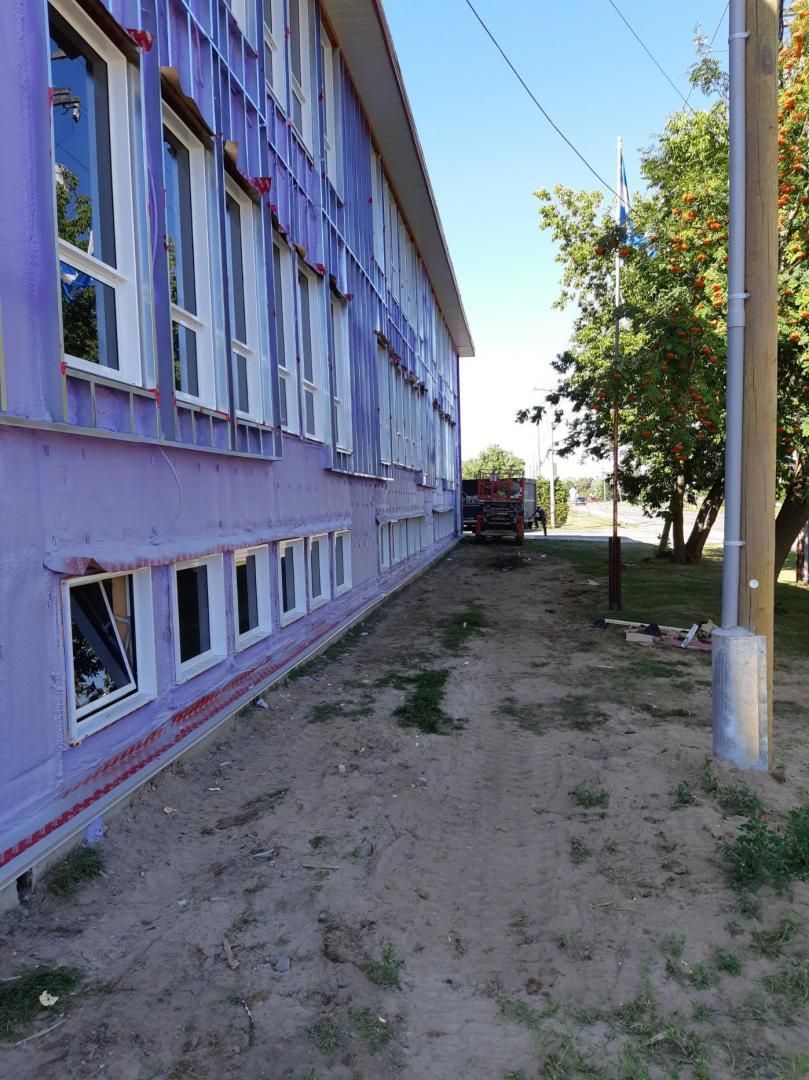 Vue latérale d'un bâtiment violet avec des fenêtres à cadre blanc, à côté d'un chemin de terre et d'arbres.