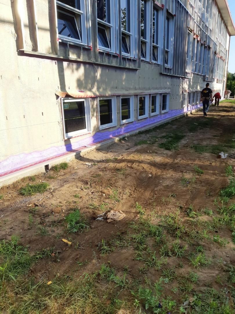 Extérieur du bâtiment avec fenêtres, isolation violette et personne marchant sur la terre et l'herbe.