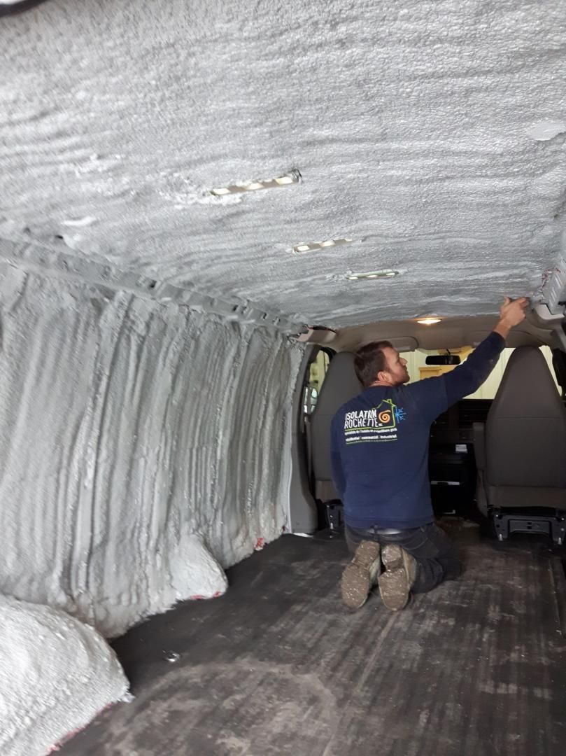 Un homme est agenouillé dans une camionnette, pulvérisant le plafond d'un isolant blanc. Intérieur noir, sol foncé.