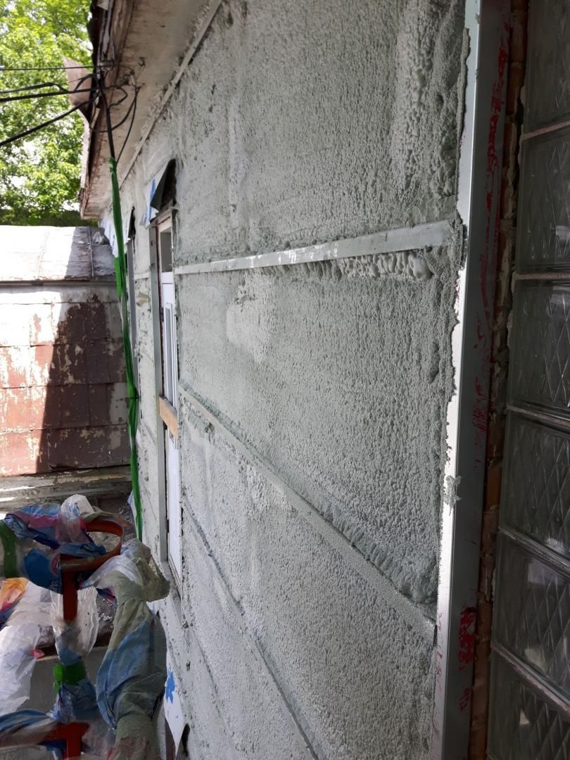 Mur extérieur avec finition en stuc et solin métallique, à côté d'une fenêtre et d'une zone encombrée.