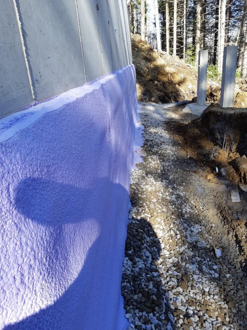 Isolation violette sur un mur de fondation en béton à côté d'une tranchée recouverte de gravier dans une zone boisée.
