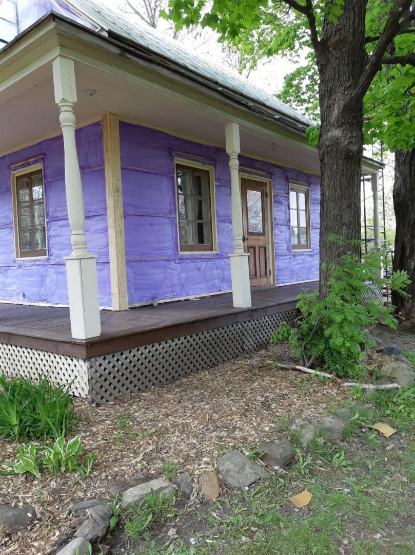 Une maison en bois peinte en lilas avec un porche, encadrée d'arbres et de verdure.