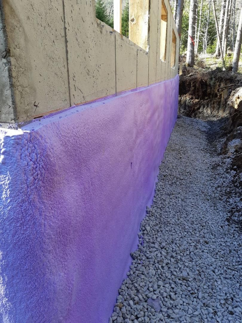 Isolation en mousse pulvérisée violette sur un mur de fondation en béton à côté de gravier ; chantier de construction.