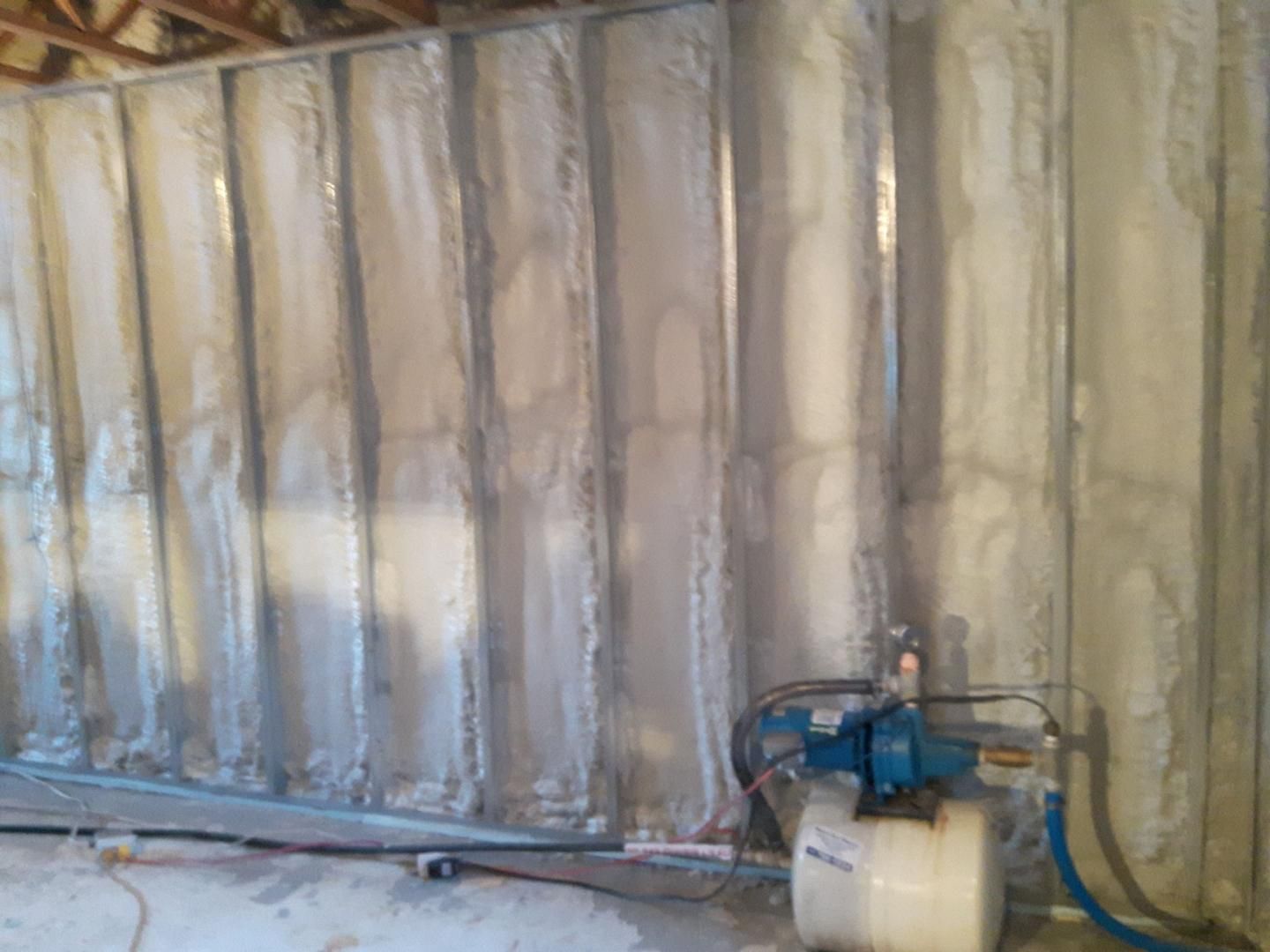 Un mur de sous-sol partiellement construit, isolé par de la mousse projetée. Au premier plan, on voit un système de pompage d'eau.