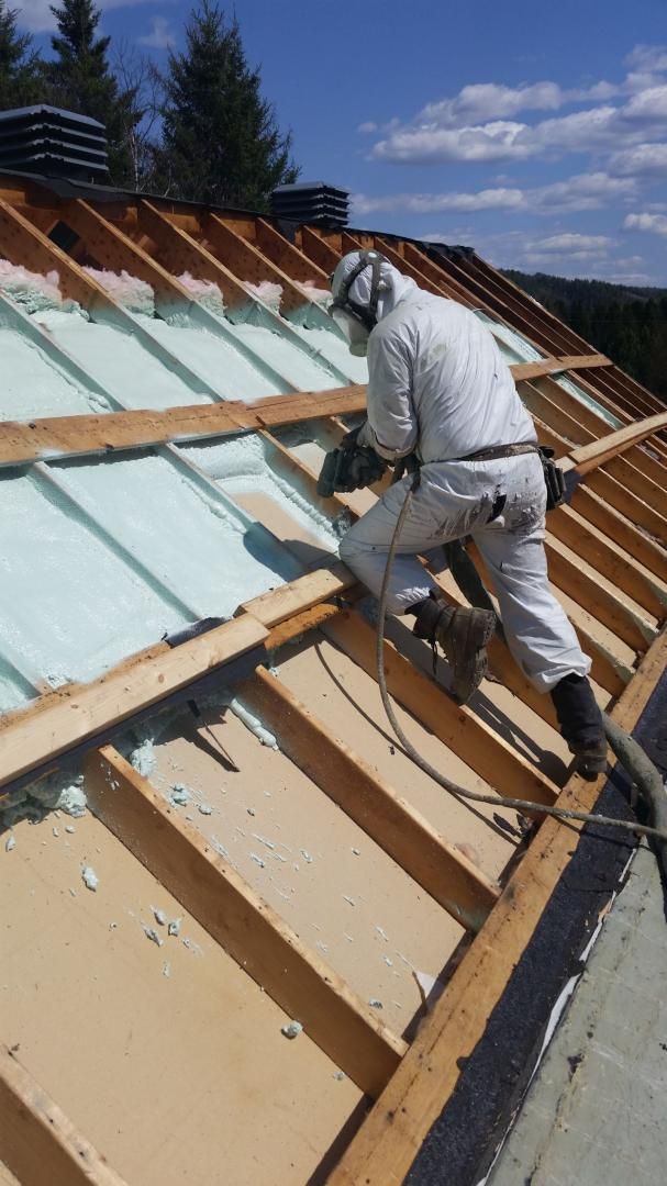Un ouvrier en costume blanc applique une isolation en mousse sur un toit en construction par une journée ensoleillée.