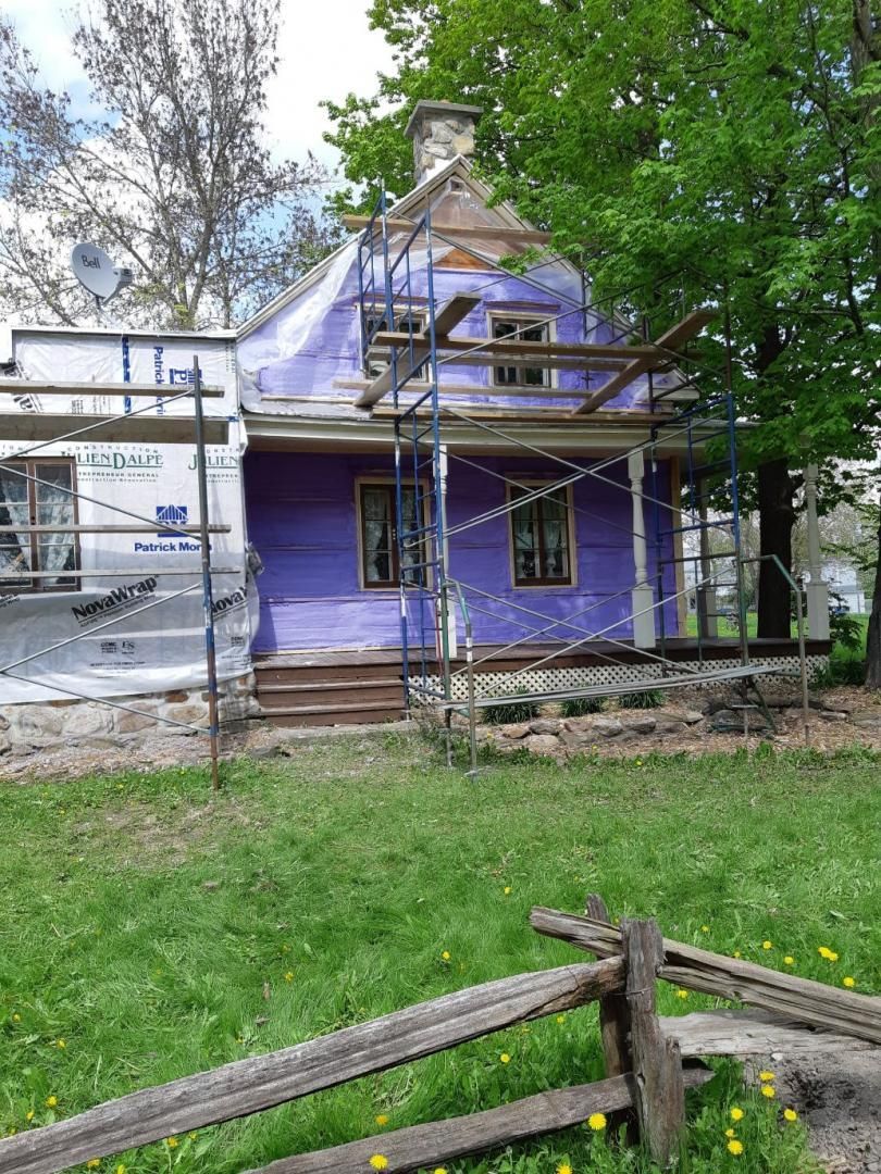 Maison en rénovation, bardage violet, échafaudage, véranda en bois, gazon vert, clôture rustique.