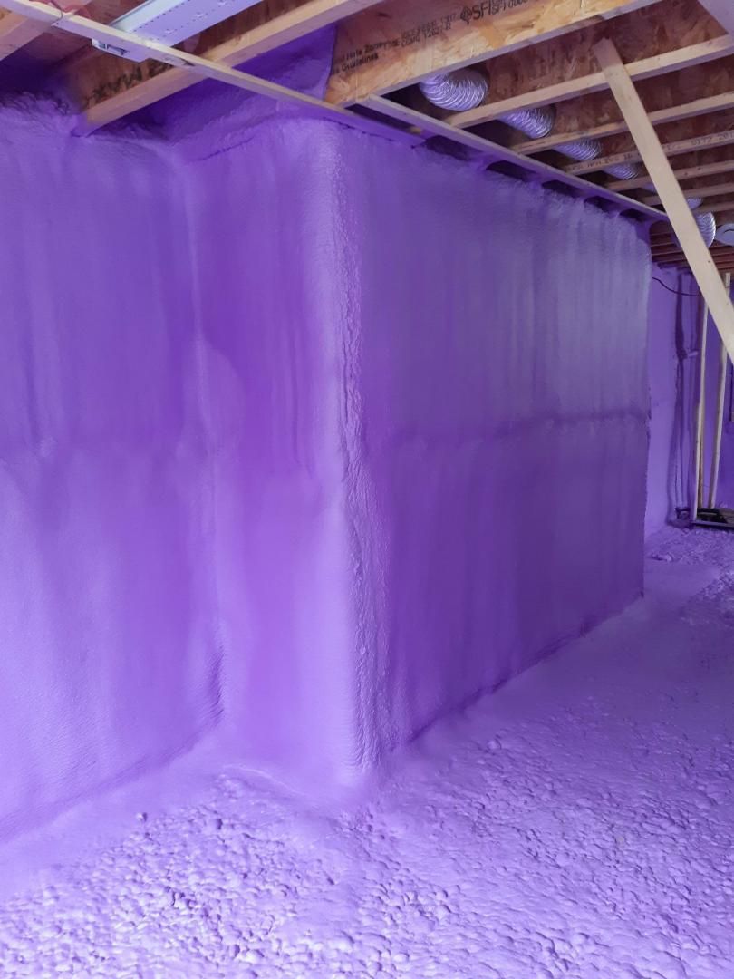 L'isolant en mousse pulvérisée violette recouvre un mur et un plancher de sous-sol, appliqué sous des poutres en bois.