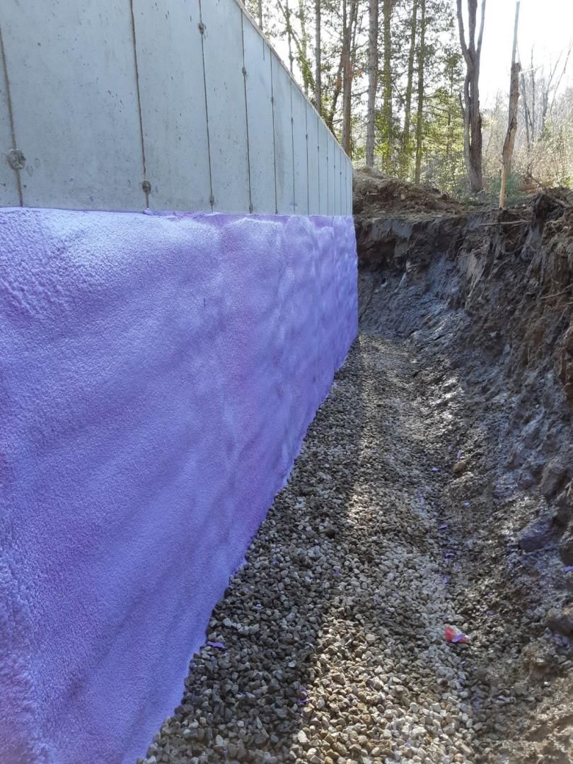 Isolation en mousse pulvérisée violette sur un mur de fondation en béton à côté d'une tranchée recouverte de gravier.