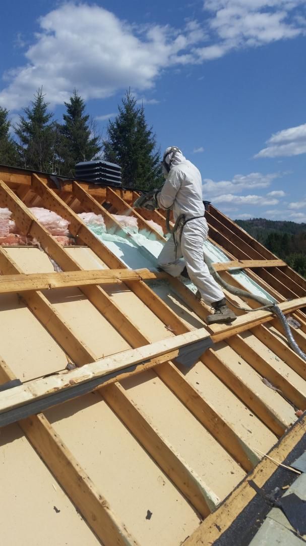 Une personne en combinaison de protection pulvérise de l'isolant sur un toit. Ciel bleu, arbres et toit partiellement isolé sont visibles.