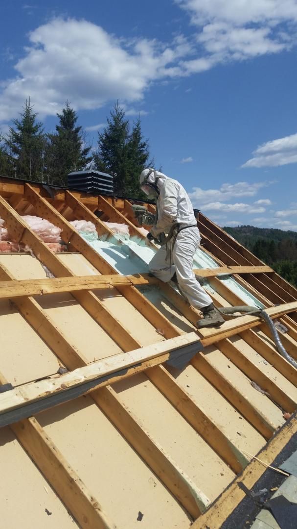 Un ouvrier en équipement de protection pulvérise de l'isolant sur un toit sous un ciel bleu.
