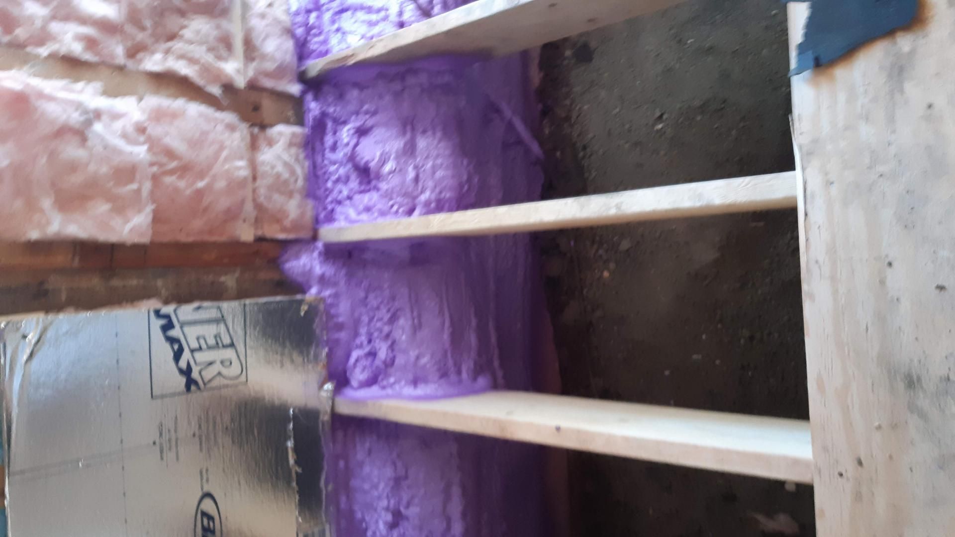 De la mousse expansive violette comble l'espace entre une étagère en bois et un mur. L'isolant et la barrière réfléchissante sont visibles.