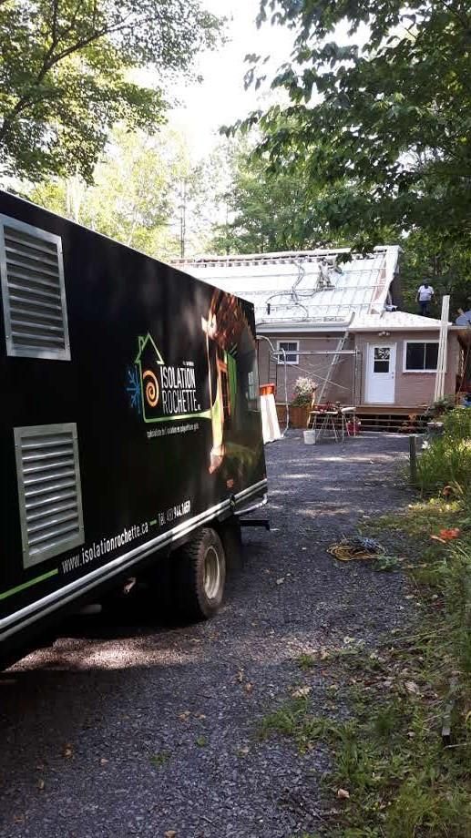 Un camion sombre garé sur du gravier, face à une petite maison en construction ; des arbres entourent la scène.
