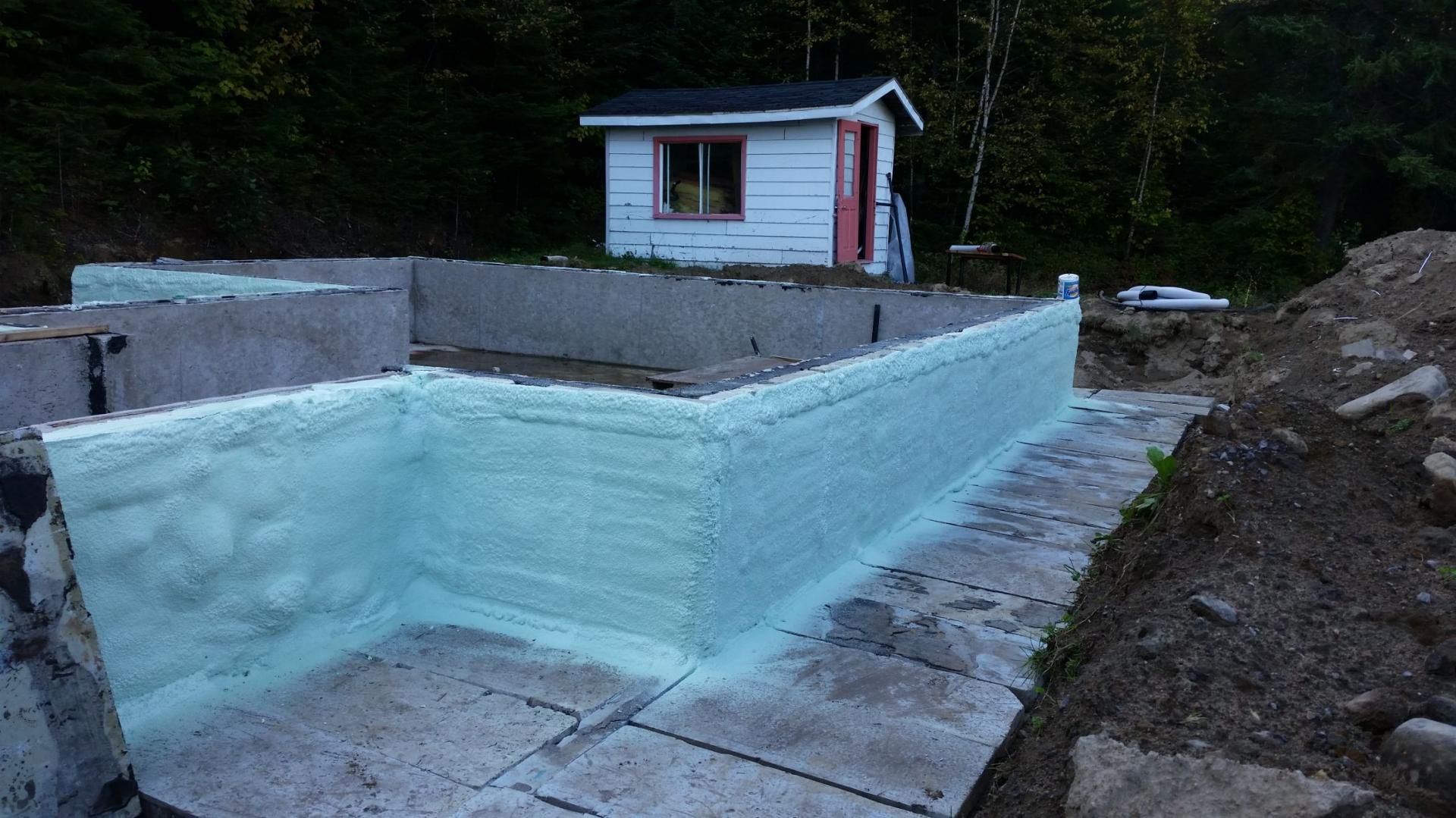 Fondation d'un bâtiment avec isolation verte en cours d'installation, petit hangar blanc en arrière-plan.