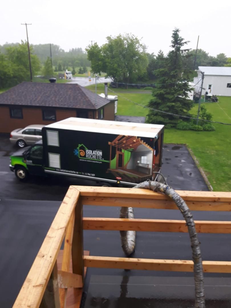 Camion avec tuyau de pompage sur un toit plat, à côté d'une structure en bois. Le camion vert porte un logo.