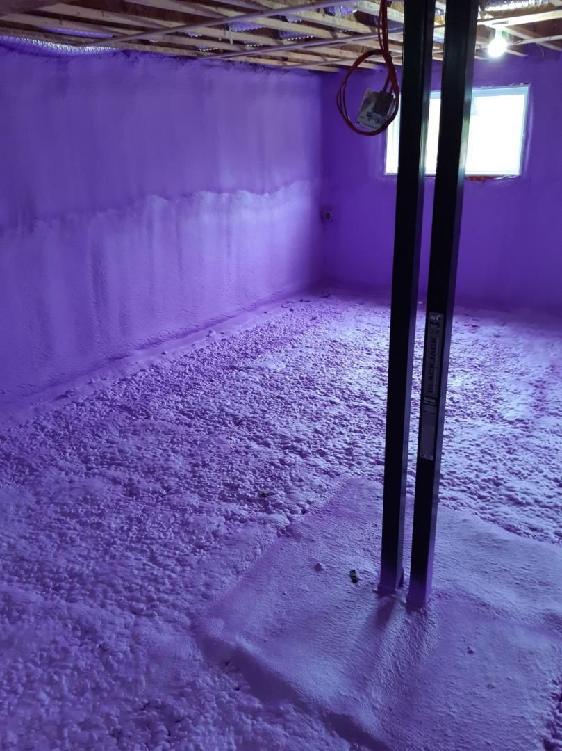 L'isolant en mousse pulvérisée violette recouvre les murs et le sol d'un sous-sol, entourant une poutre de support, avec une petite fenêtre visible.
