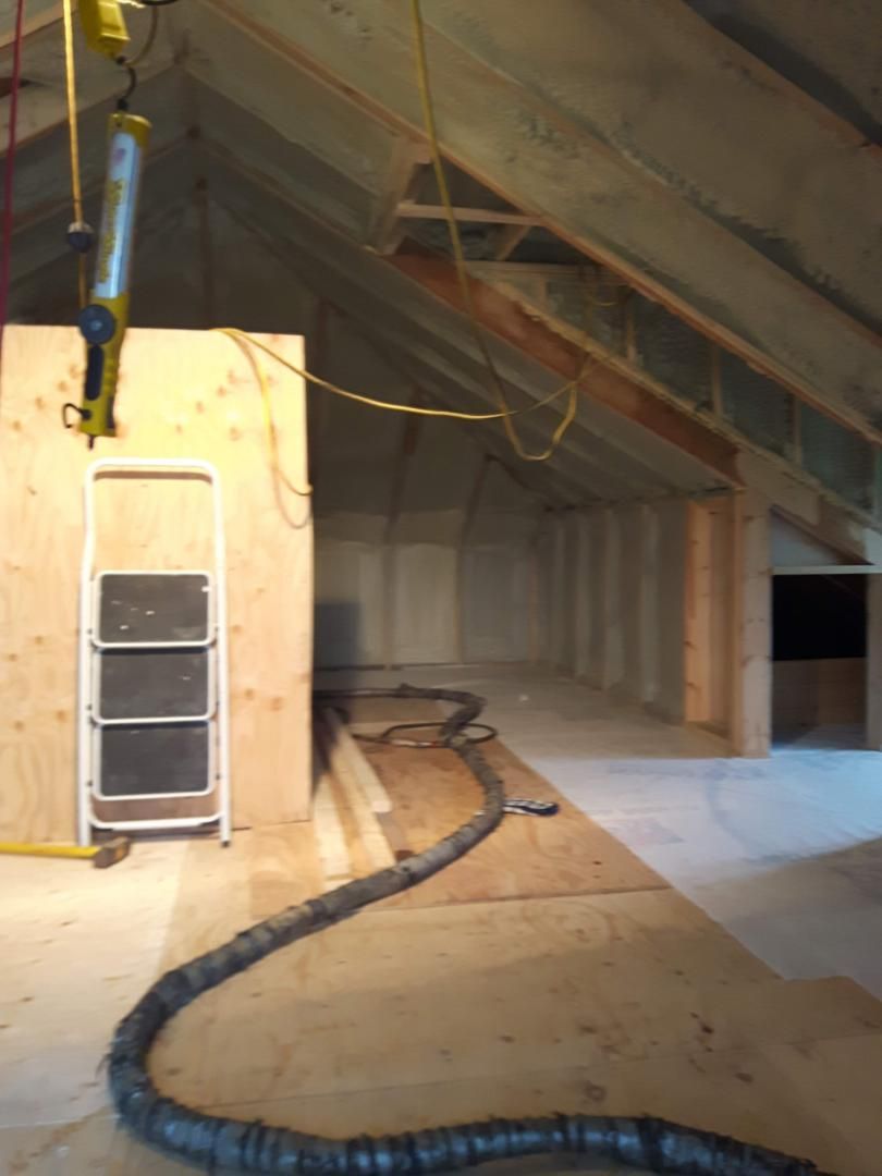 Grenier isolé avec de la mousse projetée. Tuyau gris, poutres et murs en bois. Escabeau et éclairage présents.