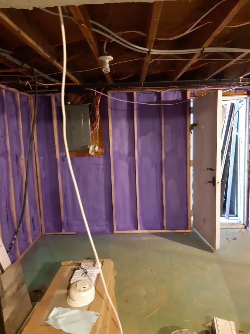 Charpente de sous-sol avec isolation violette, porte ouverte, poutres apparentes et table de travail avec objets.