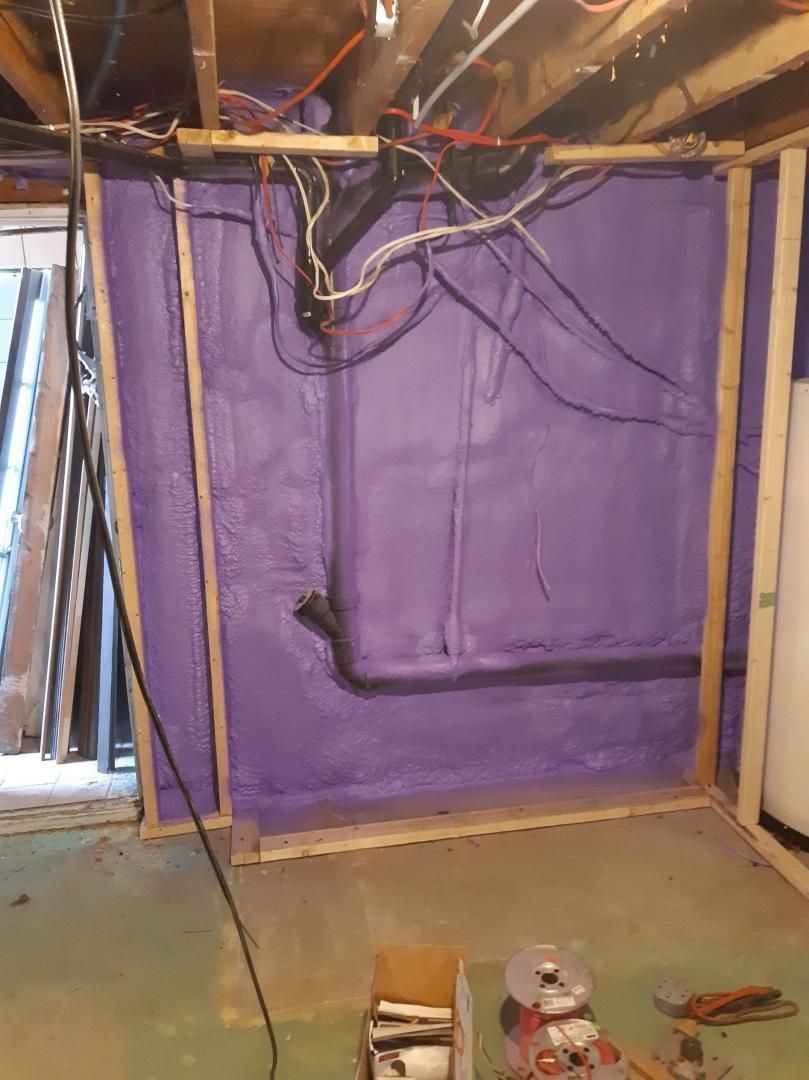Isolation en mousse pulvérisée violette sur un mur de sous-sol, encadré de bois, avec des fils et des tuyaux au-dessus.