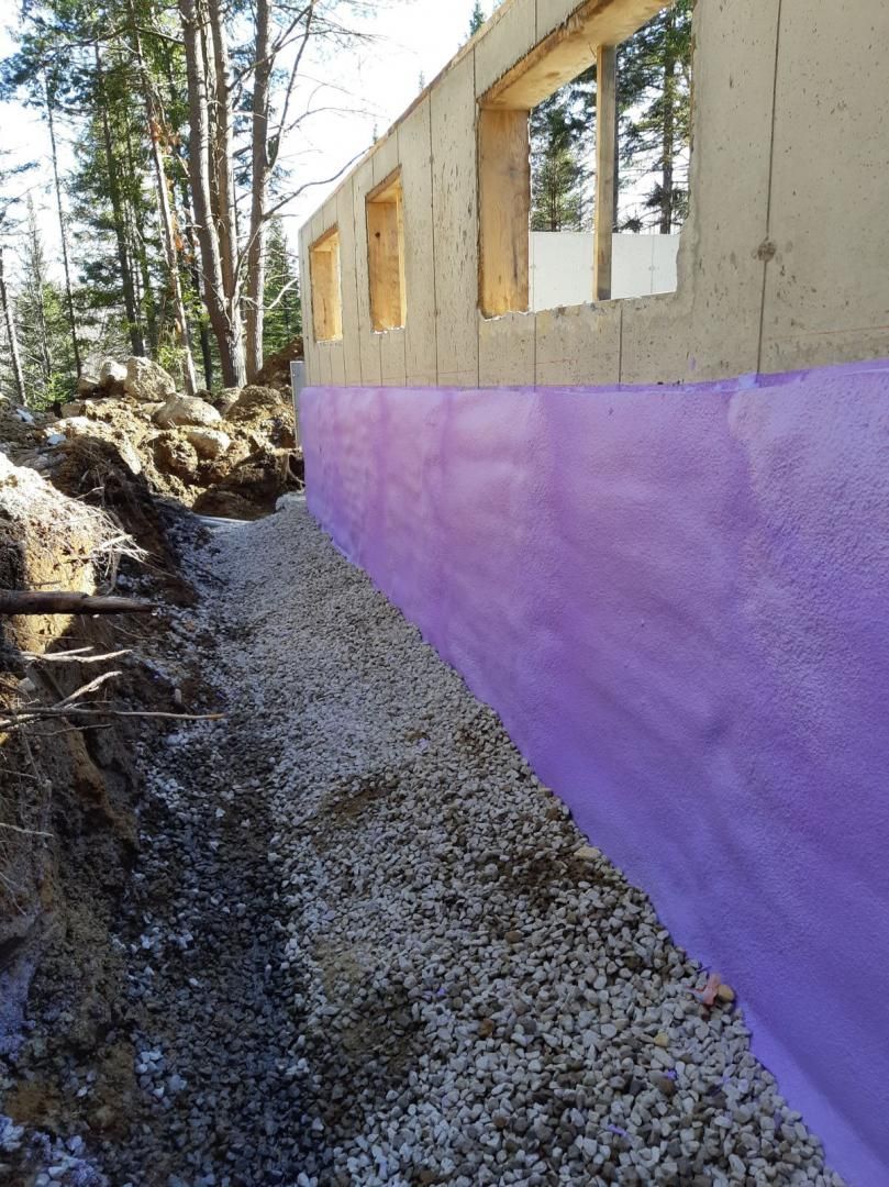 Mur de fondation recouvert de violet, à côté d'une tranchée de gravier dans une zone boisée.