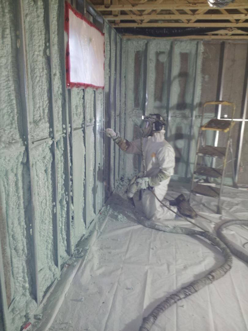 Une personne en combinaison de protection pulvérise de la mousse isolante sur un mur intérieur. Une bâche de protection blanche est posée au sol.