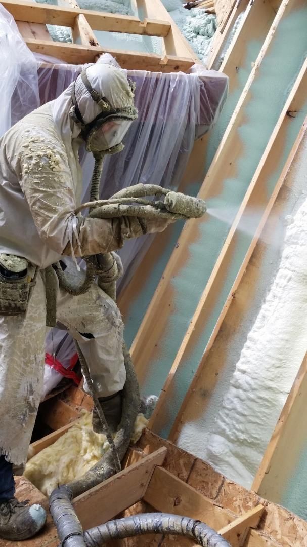 Ouvrier pulvérisant de l'isolant; portant une combinaison de protection, un masque et des gants; dans une structure de toit avec ossature en bois.