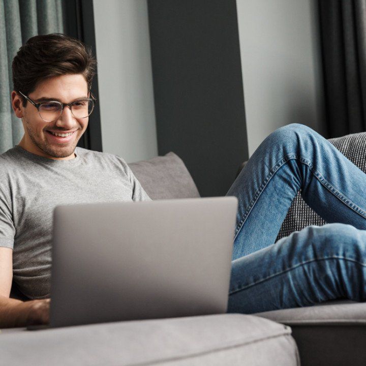 Portrait of an attractive smiling young bearded man wearing casual clothes sitting on a couch at the living room, using laptop computer