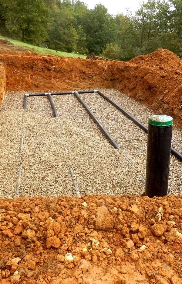 An open trench with a gravel base containing black distribution pipes and a vertical PVC pipe