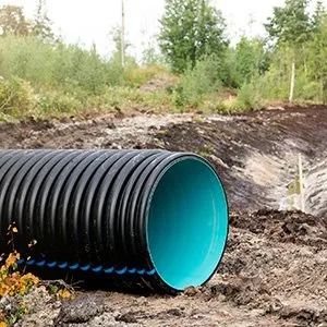 A large, black corrugated plastic drainage pipe with a bright blue interior sits on dirt near a treeline.