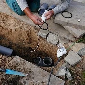A person installs a black rubber seal onto the end of a gray plastic pipe in a dirt trench.