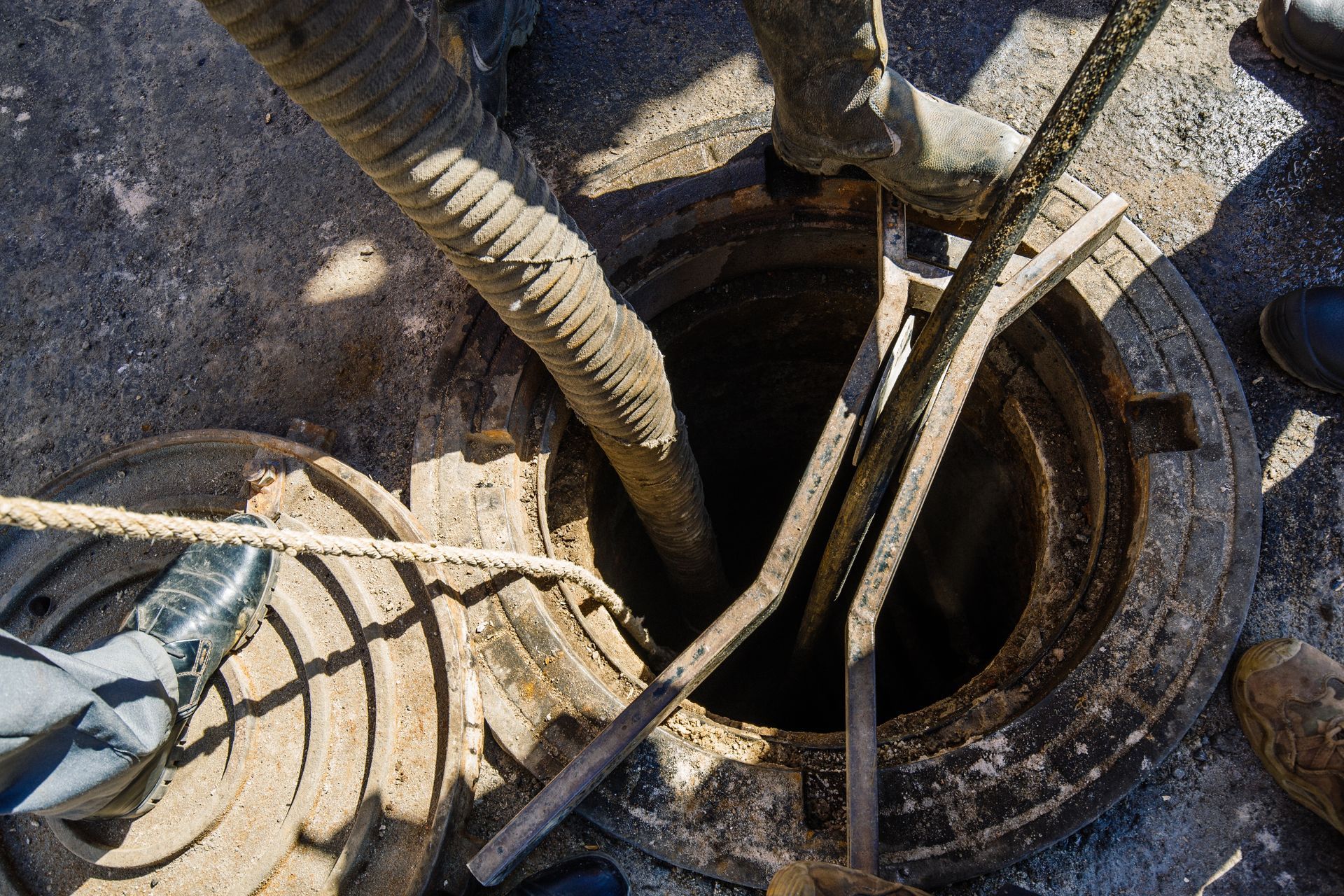 Workers use a flexible hose and metal tools to clean an open manhole on a paved street.