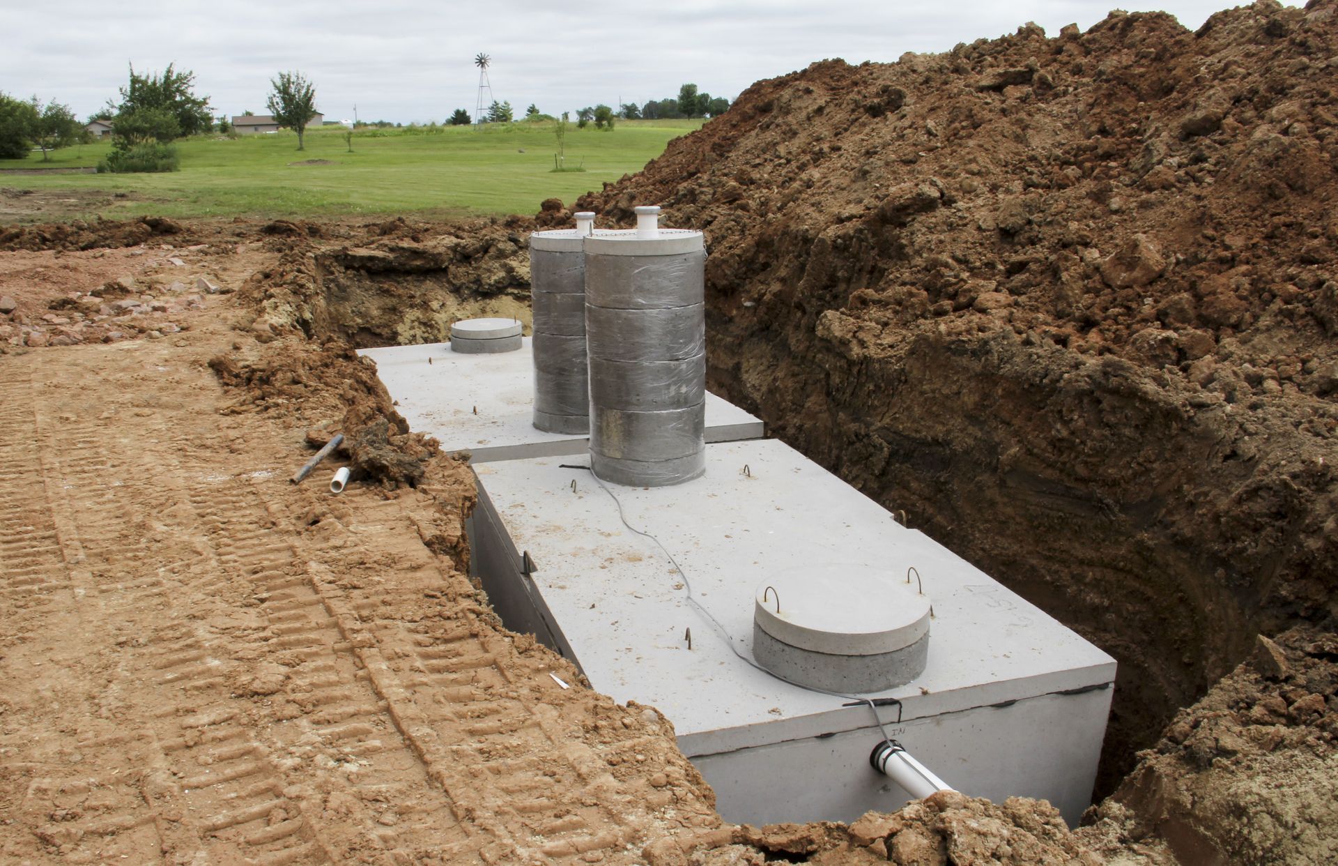 A concrete septic tank installed in a deep trench in a grassy field with piles of excavated dirt nearby.