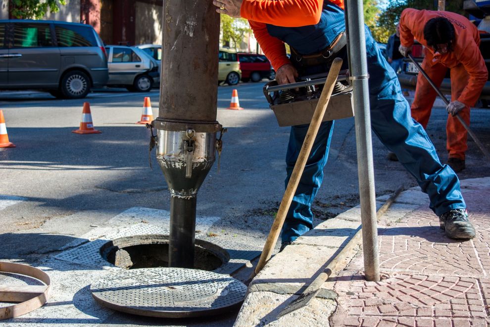 Workers in orange jumpsuits use a vacuum truck hose to clean a manhole in a street lined with safety cones.