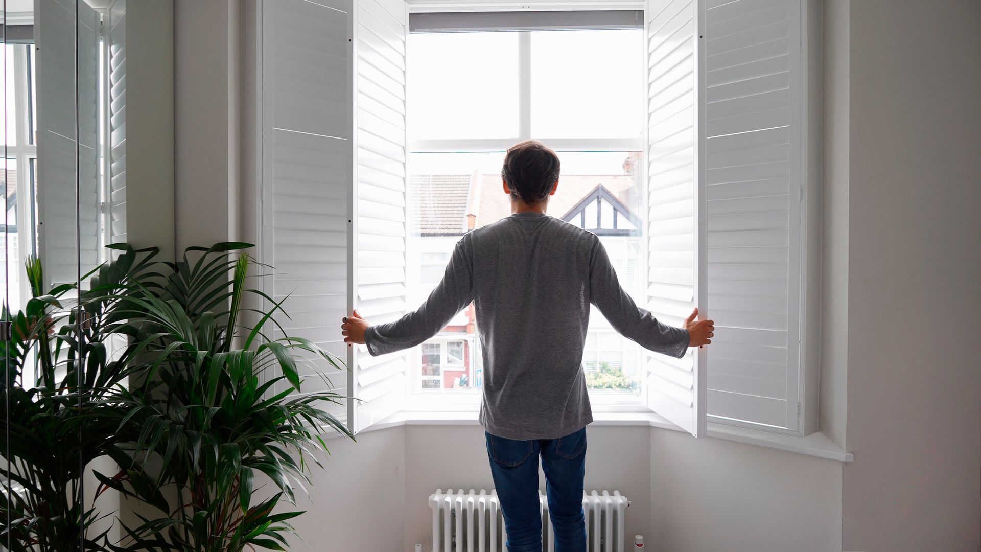 Un uomo apre le persiane bianche della finestra e guarda fuori, in una stanza luminosa.