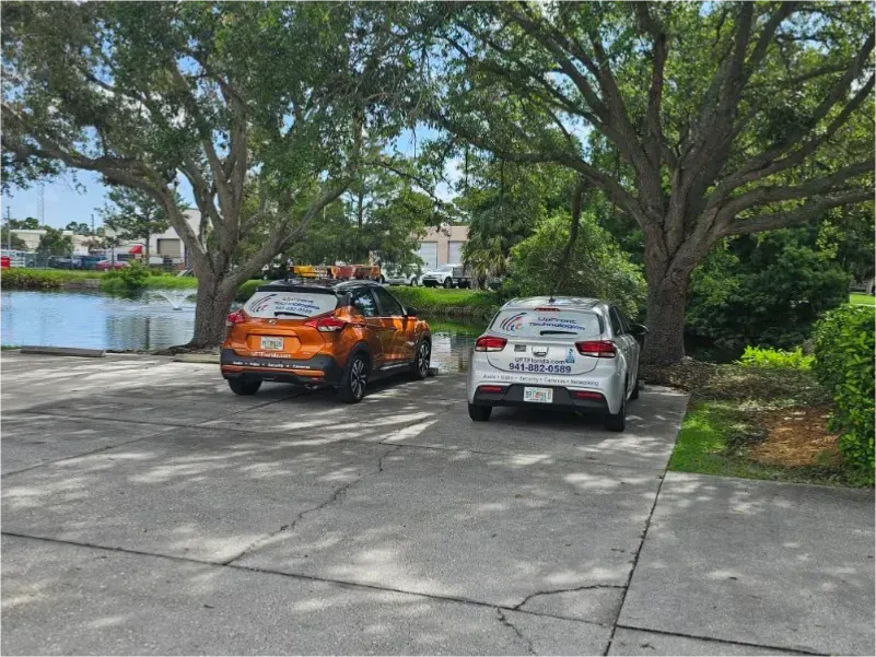 Two cars parked on concrete by a body of water, under trees. One orange, one silver.
