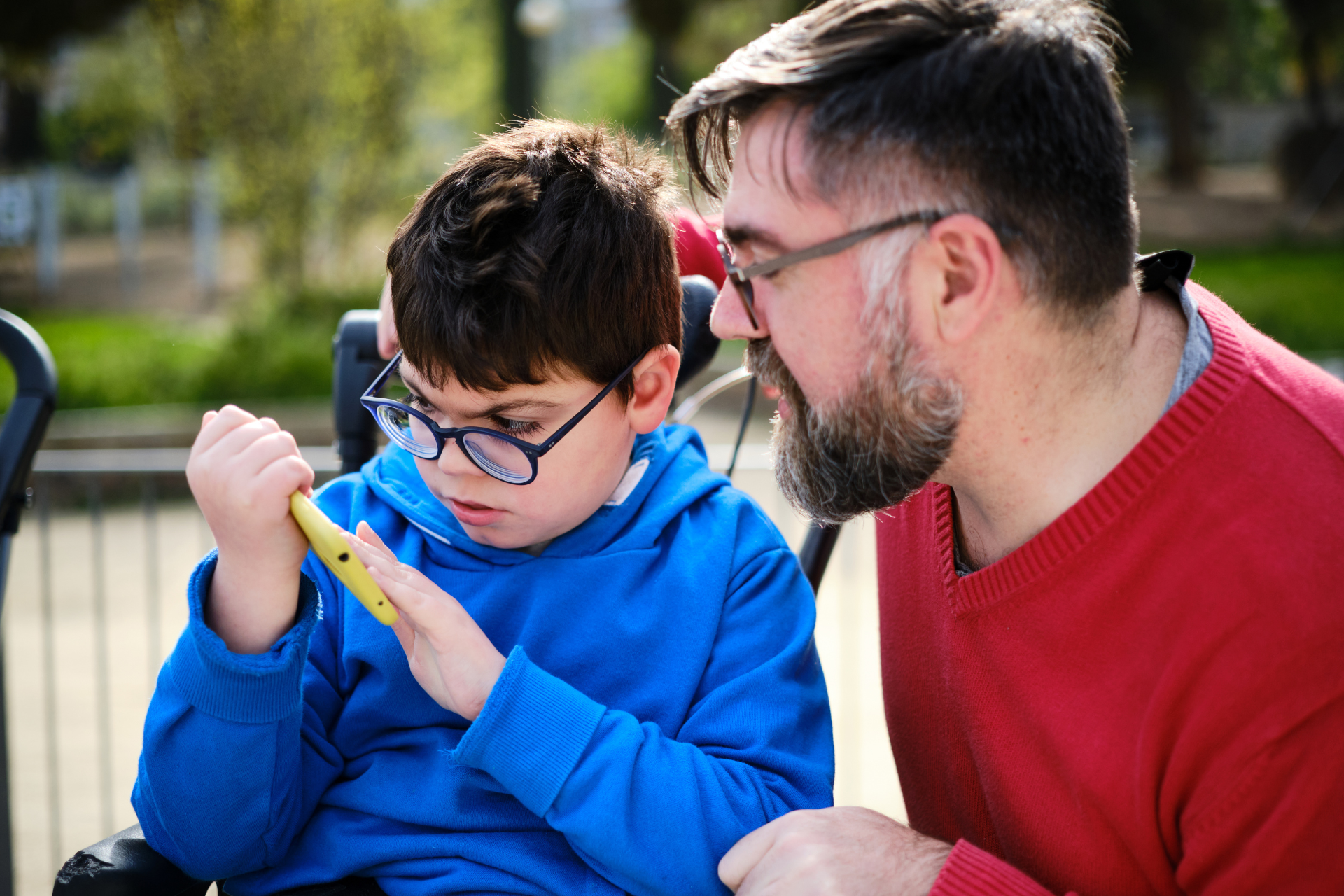 A man and a boy in a wheelchair are looking at a cell phone.