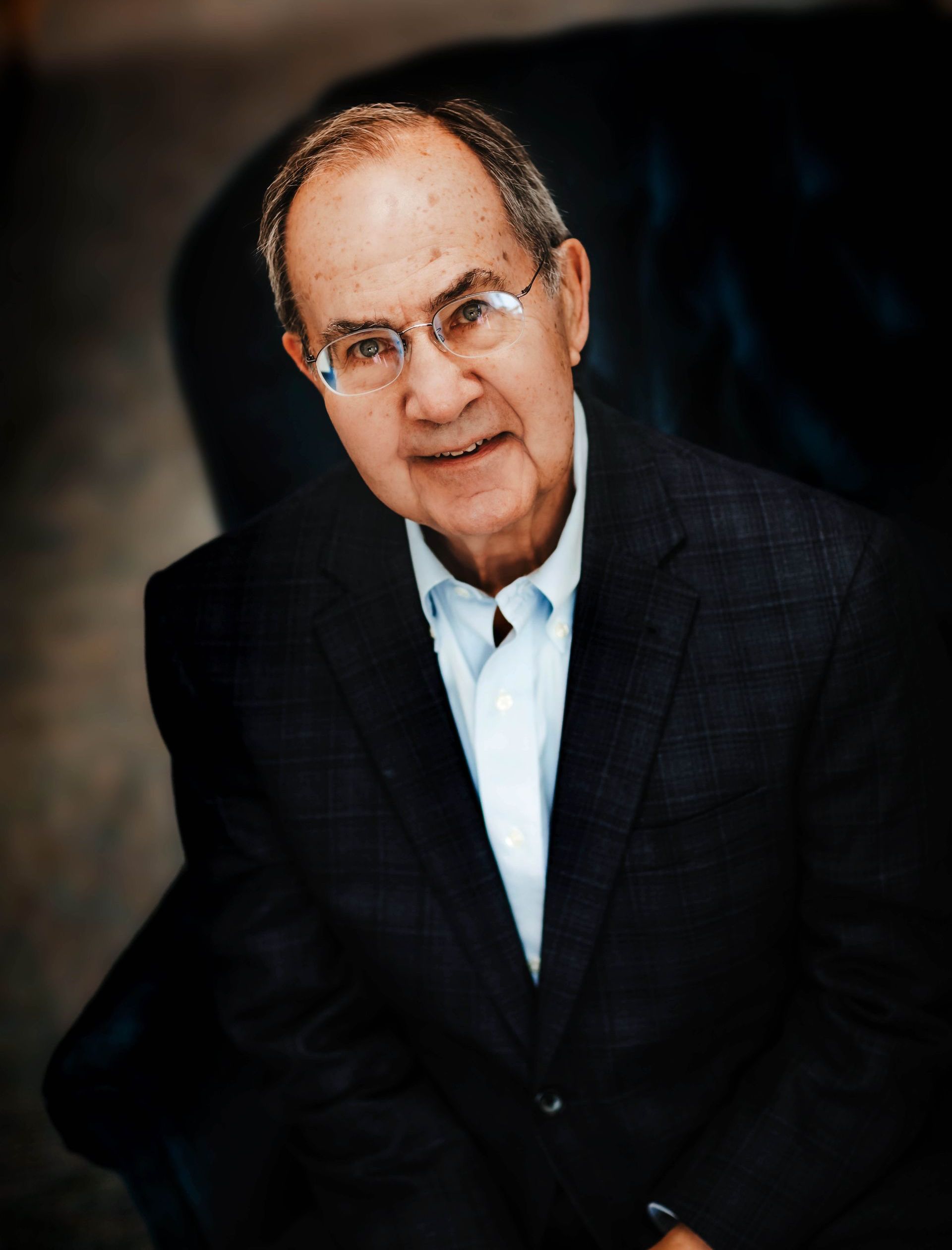 A man wearing glasses and a suit is sitting in a chair.