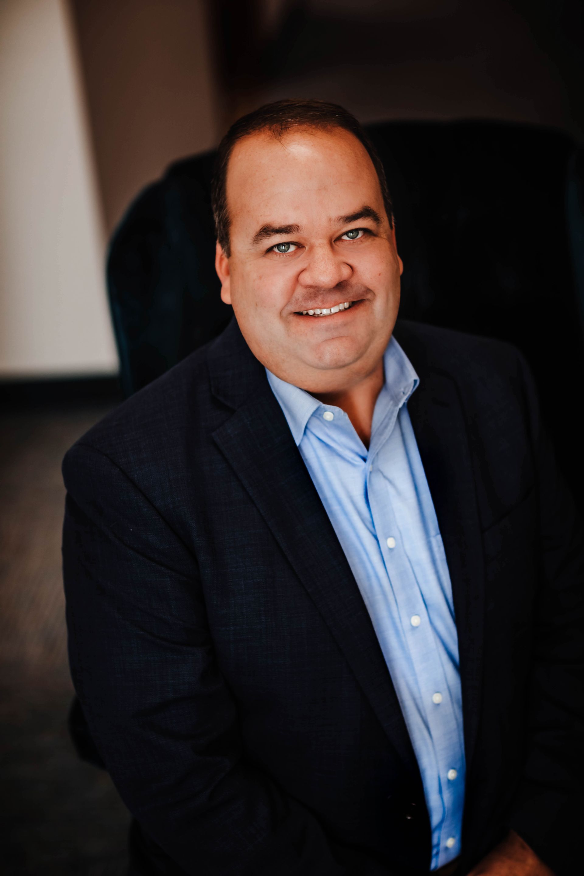 A man in a suit and blue shirt is sitting in a chair and smiling.