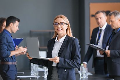 Woman in a blazer holds laptop, smiles at the camera in a modern office, colleagues in background.