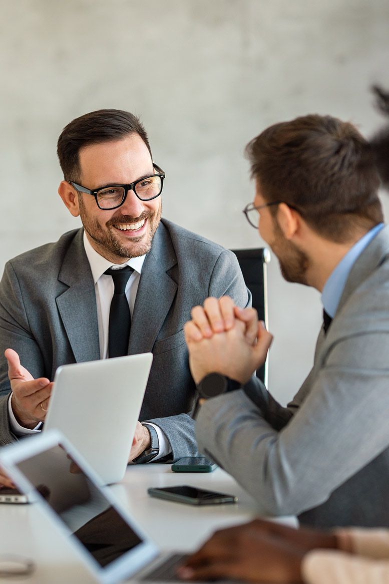 Two men in suits, smiling, discussing at a table with laptops.