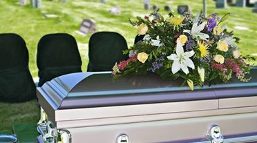 A casket with floral arrangement at a funeral, set in a grassy area with chairs.