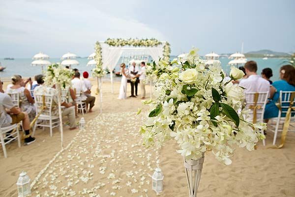 Beach wedding ceremony with white flowers, arch, guests, and ocean backdrop.