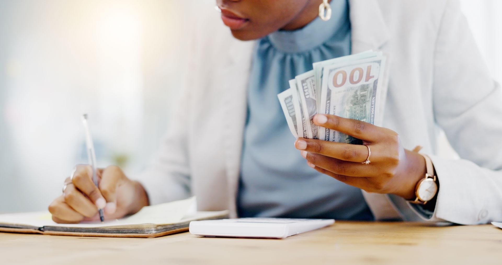 A woman is sitting at a table holding a bunch of money and writing in a notebook.