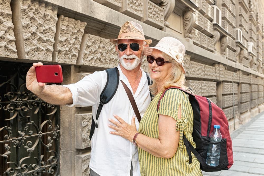 Casal tirando uma selfie ao ar livre; homem de camisa branca e chapéu, mulher de blusa amarela e chapéu; prédio ao fundo.