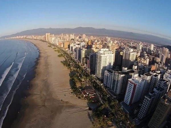 Vista aérea de uma cidade costeira com uma longa praia de areia, edifícios e montanhas ao fundo, sob um céu azul.