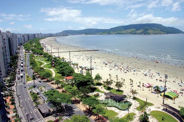 Orla da praia de Santos com muita gente, prédios, estrada, árvores e uma montanha sob um céu azul.